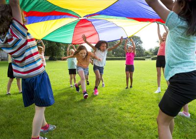 A group of children playing outdoors on a grassy field, lifting a large, colorful play parachute overhead while two kids run excitedly underneath it.