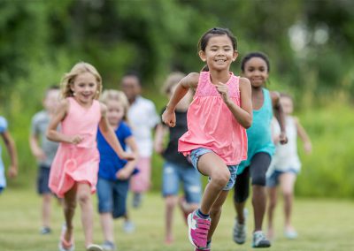 A group of happy young children running towards the camera across a grassy field on a sunny day, led by a girl in a pink top.