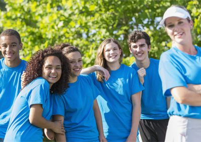 A cheerful group of teenagers wearing matching blue t-shirts smiling together outdoors in front of lush green trees, with an adult leader in a white visor blurred in the foreground.
