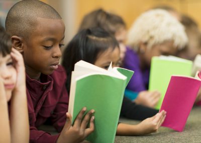 A line of young children lying on the floor, focused on reading colorful books together in a classroom or library setting.